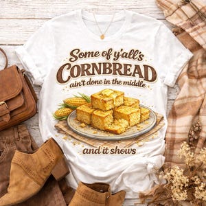 Puede incluir: Camiseta blanca con el texto "Some of y'all's CORNBREAD ain't done in the middle and it shows." La camiseta presenta un gráfico de cuadrados de pan de maíz con mantequilla, maíz y un plato. Forma parte de una colección temática de comida.