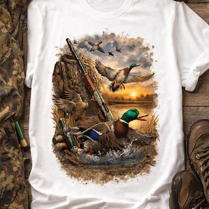 Puede incluir: Camiseta blanca con una ilustración detallada de patos en vuelo sobre el agua, una escopeta y un fondo de puesta de sol. El diseño incluye patos, agua y una escena de paisaje, con elementos de caza.