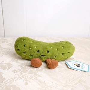 May include: A plush green pickle toy with black dot details and a smiling face. Two small, brown corduroy-like objects are next to the pickle. A tag with a cartoon cat logo is attached.