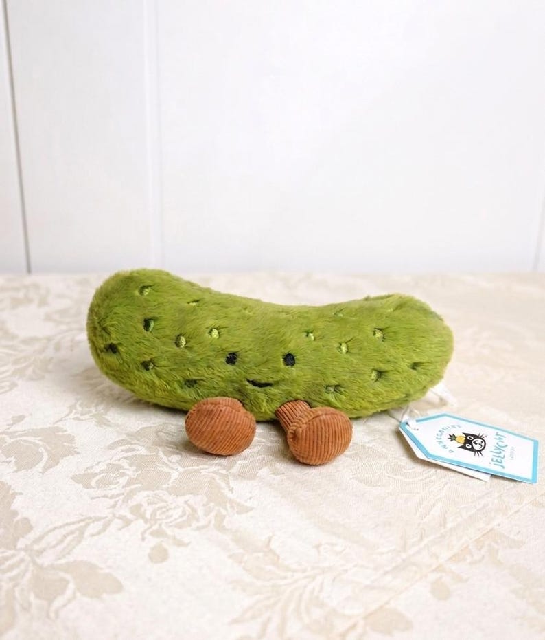 May include: A plush green pickle toy with black dot details and a smiling face. Two small, brown corduroy-like objects are next to the pickle. A tag with a cartoon cat logo is attached.