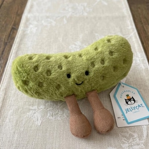 May include: A plush, green pickle toy with a smiling face and two brown legs. The toy has a textured surface with small, round indentations. A tag with the Jellycat logo is attached.