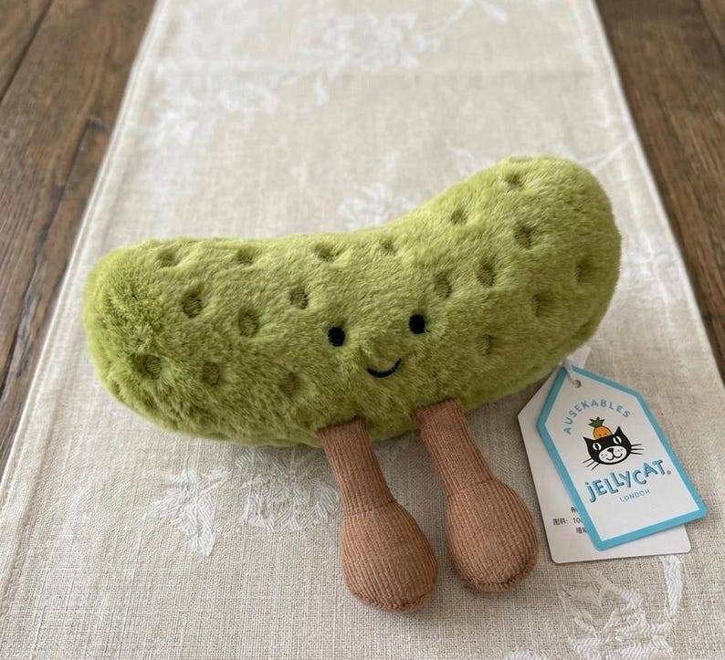 May include: A plush, green pickle toy with a smiling face and two brown legs. The toy has a textured surface with small, round indentations. A tag with the Jellycat logo is attached.