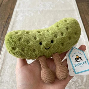 May include: A plush green pickle toy with a smiling face and brown corduroy legs. The toy has a textured surface with small, dark indentations. A tag with a black cat logo is attached.