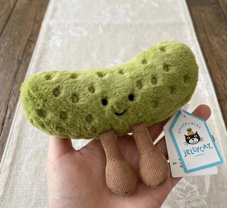 May include: A plush green pickle toy with a smiling face and brown corduroy legs. The toy has a textured surface with small, dark indentations. A tag with a black cat logo is attached.