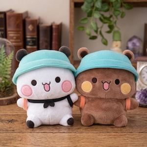 May include: Two plush bear dolls, one white and one brown, each wearing a light blue hat. The white bear has a black bow tie and the brown bear has orange straps. Both have pink cheeks and are smiling.