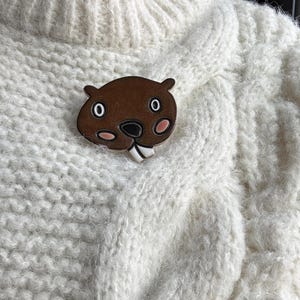 Könnte beinhalten: Ein brauner Biber-Emaille-Ansteckpin mit weißen Zähnen, rosa Wangen und schwarzen Augen ist an einem cremefarbenen Zopfstrickpullover befestigt. Der Pin ist ein skurriles Accessoire, das dem Pullover eine persönliche Note verleiht.