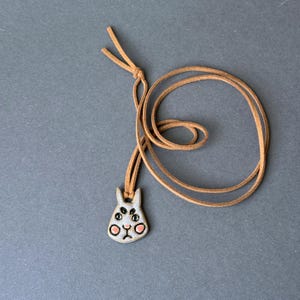Op de afbeelding: Handgemaakte keramische hangerketting met een grillig konijnontwerp. De hanger is lichtgrijs met zwarte en roze accenten, geregen op een bruin leren koord. Het konijn heeft meerdere ogen en een kleine roze mond. Op een grijze achtergrond.