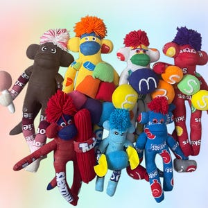 Puede incluir: Una colección de ocho monos de calcetín hechos a mano con diversos patrones de tela coloridos, incluyendo estampados de dulces como Skittles y Airheads. Cada mono presenta un pompón de lana en la cabeza y expresiones faciales distintas.