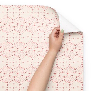 Puede incluir: Rollo de papel pintado con un patrón repetitivo de pequeños corazones rosas sobre un fondo crema. El patrón forma rombos. Una mano levanta una esquina del papel pintado, revelando el reverso blanco.