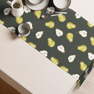 Pode incluir: Um caminho de mesa cinza escuro com um padrão repetido de peras amarelas e metades de pera. São visíveis utensílios de mesa de cerâmica branca, incluindo um jarro, xícaras e pratos. Uma colher e um garfo também estão presentes.