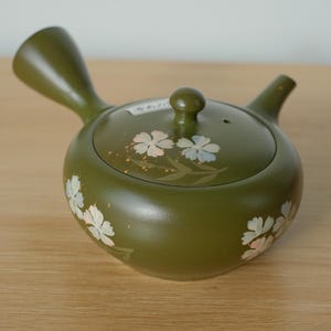 Può includere: Teiera in ceramica verde oliva con manico laterale e beccuccio piccolo. Il coperchio è ornato da un piccolo pomello e disegni floreali bianchi e rosa. La teiera è appoggiata su una superficie di legno.