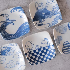Peut inclure: Ensemble de six assiettes carrées en céramique avec des illustrations bleues et blanches. Chaque assiette présente un design différent, dont Snoopy surfant, le mont Fuji et des motifs de nuages. Les assiettes ont une esthétique japonaise.