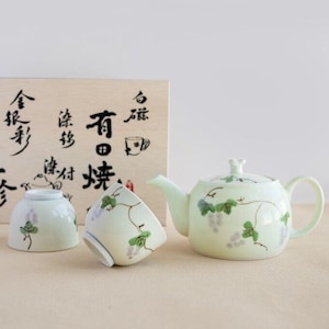 以下が含まれることがあります： 薄緑色の陶器のティーセット。ティーポット、カップ2つ、木箱が含まれています。緑の葉と紫のブドウのブドウの木の模様が特徴です。木箱には日本語の文字が書かれています。