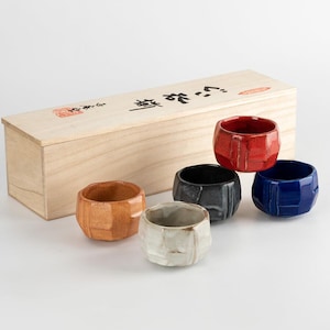 May include: A set of six ceramic sake cups in various colors, including red, blue, black, and brown. The cups have a faceted design and are displayed with a wooden box. The box has Japanese text and a red stamp.