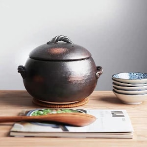 Könnte beinhalten: Ein dunkelbrauner Keramikkochtopf mit Deckel und gedrehtem Griff. Der Topf steht auf einer gewebten Matte, daneben kleine Schalen und ein Holzlöffel. Im Vordergrund ist eine Zeitschrift zu sehen.