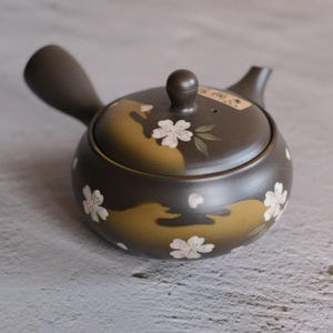 Puede incluir: Tetera de cerámica marrón con asa lateral, boquilla y tapa. La tetera está decorada con diseños de flores de cerezo blancas y hojas verdes. El fondo de la tetera es una mezcla de marrón y dorado.