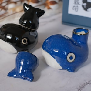 Puede incluir: Colección de objetos de cerámica con forma de ballena. Uno es negro con vientre blanco, otro azul con vientre blanco, y un tercero es una pequeña cola azul. Cada uno tiene un detalle de ojo circular.