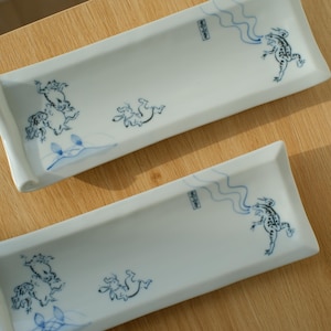 Könnte beinhalten: Zwei rechteckige weiße Keramik-Servierplatten mit blauen Illustrationen von Fröschen und anderen Kreaturen. Die Platten haben leicht gebogene Kanten und liegen auf einer hellen Holzoberfläche.