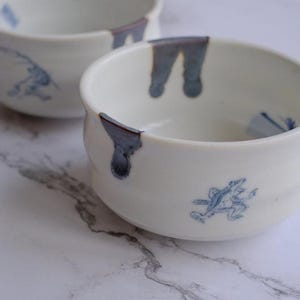May include: Two white ceramic bowls with blue accents and frog illustrations. The bowls have a rounded shape with a slightly flared rim. The blue accents are on the rim and the frog illustrations are on the side of the bowls.