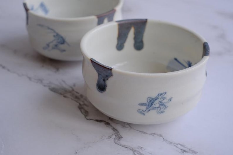 May include: Two white ceramic bowls with blue accents and frog illustrations. The bowls have a rounded shape with a slightly flared rim. The blue accents are on the rim and the frog illustrations are on the side of the bowls.