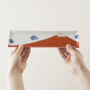 May include: Rectangular ceramic serving dish featuring a Mount Fuji design. The mountain is red with a white, snow-covered peak. Blue clouds are painted on the white background. The dish is held by two hands.