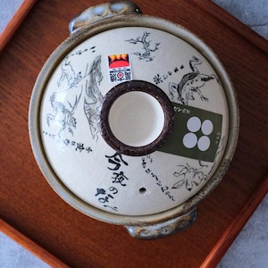 Puede incluir: Una olla japonesa donabe de color crema con una tapa que presenta ilustraciones negras de ranas y plantas. La tapa tiene un asa marr&oacute;n y una peque&ntilde;a abertura redonda. La olla est&aacute; sobre una bandeja de madera.