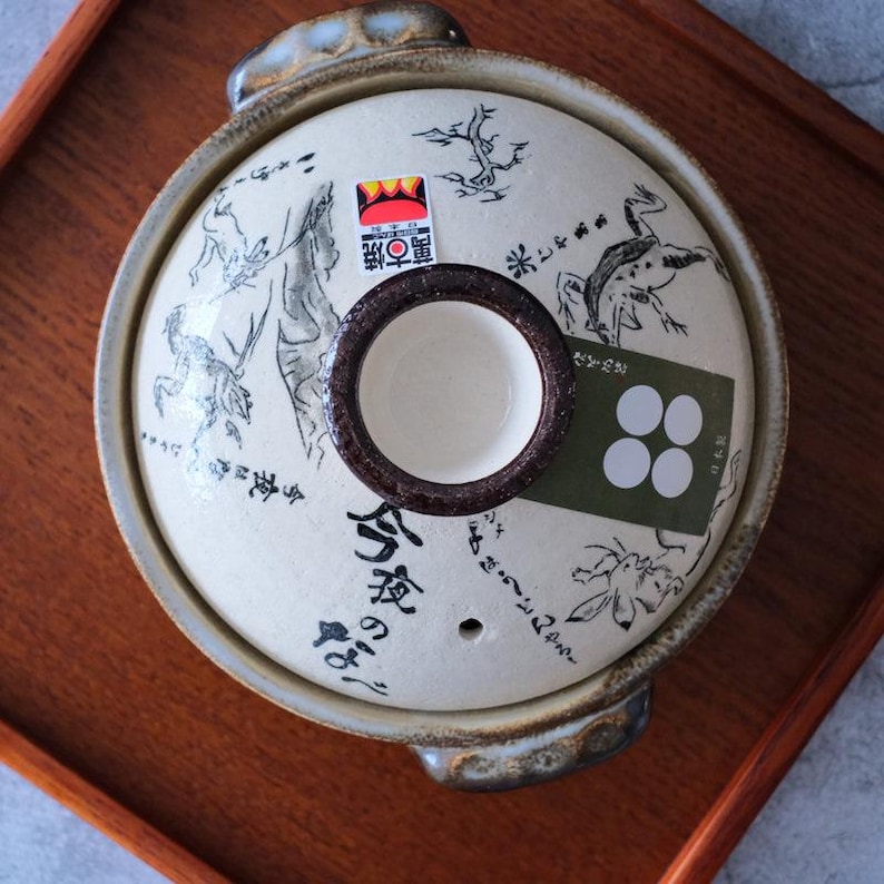 Puede incluir: Una olla japonesa donabe de color crema con una tapa que presenta ilustraciones negras de ranas y plantas. La tapa tiene un asa marr&oacute;n y una peque&ntilde;a abertura redonda. La olla est&aacute; sobre una bandeja de madera.
