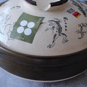 Puede incluir: Una olla japonesa donabe de cer&aacute;mica con tapa. La olla es de color marr&oacute;n oscuro, y la tapa es beige con ilustraciones negras de una rana, un &aacute;rbol y otros dise&ntilde;os. Una pegatina verde con c&iacute;rculos blancos est&aacute; en la tapa.