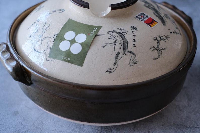 Puede incluir: Una olla japonesa donabe de cer&aacute;mica con tapa. La olla es de color marr&oacute;n oscuro, y la tapa es beige con ilustraciones negras de una rana, un &aacute;rbol y otros dise&ntilde;os. Una pegatina verde con c&iacute;rculos blancos est&aacute; en la tapa.