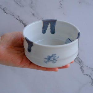 May include: A white ceramic bowl with a rounded shape, featuring dark blue drips around the rim and a blue floral design on the side. The bowl is held in a hand.
