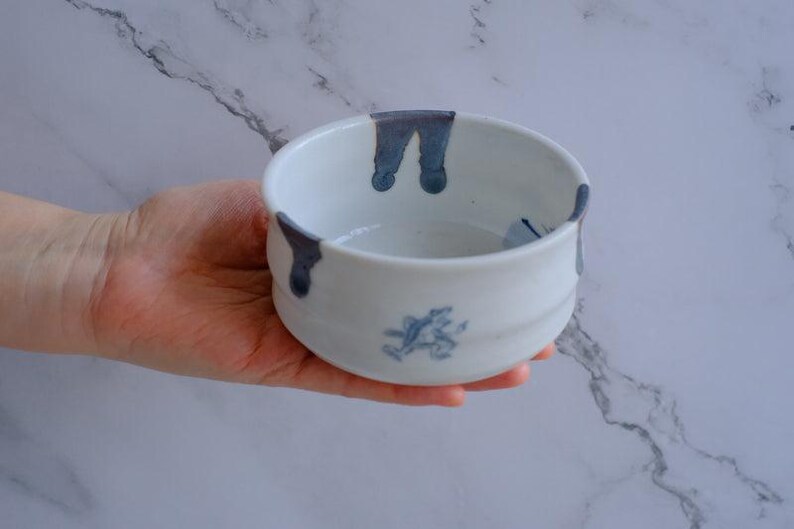 May include: A white ceramic bowl with a rounded shape, featuring dark blue drips around the rim and a blue floral design on the side. The bowl is held in a hand.