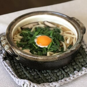 Puede incluir: Un cuenco de fideos udon con huevo escalfado, verduras verdes y champi&ntilde;ones. El cuenco de cer&aacute;mica tiene un exterior marr&oacute;n oscuro y est&aacute; sobre un salvamanteles de ganchillo gris y blanco. La yema es de color naranja brillante.