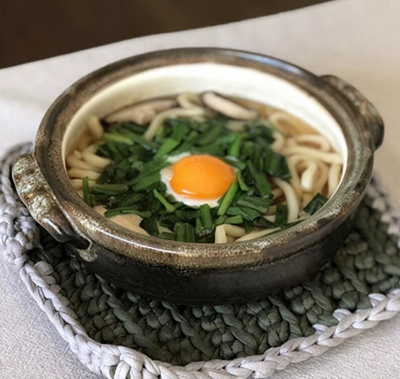 Puede incluir: Un cuenco de fideos udon con huevo escalfado, verduras verdes y champi&ntilde;ones. El cuenco de cer&aacute;mica tiene un exterior marr&oacute;n oscuro y est&aacute; sobre un salvamanteles de ganchillo gris y blanco. La yema es de color naranja brillante.