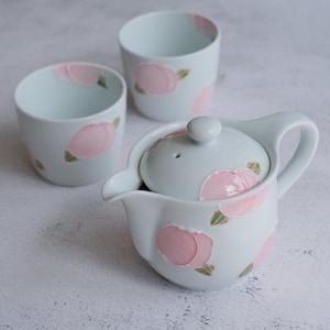 May include: A light blue ceramic tea set with pink floral designs and green leaves. The set includes a teapot with a lid and two cups. The teapot has a handle and spout. The cups and teapot are decorated with pink flowers.