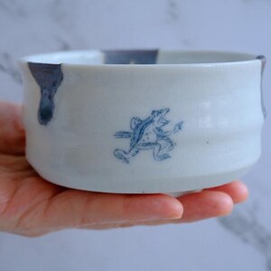 May include: White ceramic bowl with a blue frog illustration. The bowl has a rounded shape with a slightly flared rim and a blue accent on the edge. The frog is depicted in a running pose, adding a whimsical touch to the design.
