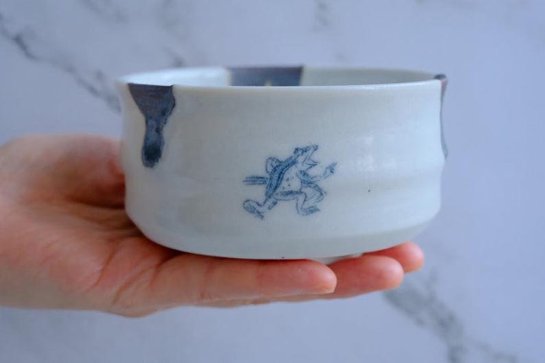 May include: White ceramic bowl with a blue frog illustration. The bowl has a rounded shape with a slightly flared rim and a blue accent on the edge. The frog is depicted in a running pose, adding a whimsical touch to the design.