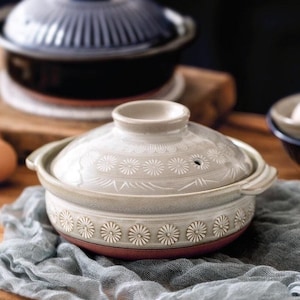 May include: A ceramic cooking pot with a lid. The pot is light gray with a red base and a floral pattern around the rim and on the lid. The lid has a small vent hole and a handle. Another pot is visible in the background.