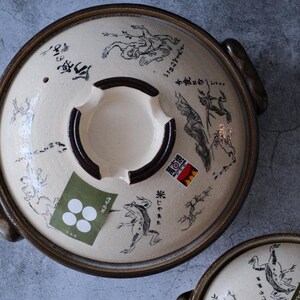 Puede incluir: Tapa de olla japonesa donabe de cer&aacute;mica color crema con borde y asas marrones oscuros. La tapa presenta ilustraciones negras de ranas y otras figuras, junto con texto y pegatinas japonesas.