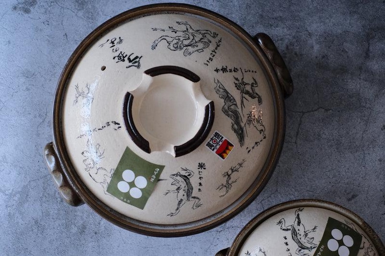 Puede incluir: Tapa de olla japonesa donabe de cer&aacute;mica color crema con borde y asas marrones oscuros. La tapa presenta ilustraciones negras de ranas y otras figuras, junto con texto y pegatinas japonesas.