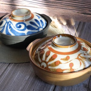May include: Two ceramic Japanese donabe pots with lids. One pot is dark grey with a blue and white swirl design on the lid. The other is tan with an orange and white floral pattern on the lid. Both have handles.