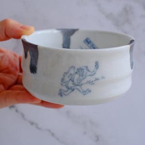 May include: White ceramic bowl with a blue illustration of a running figure. The rim has blue paint drips. The bowl has a slightly textured surface and a rounded shape.