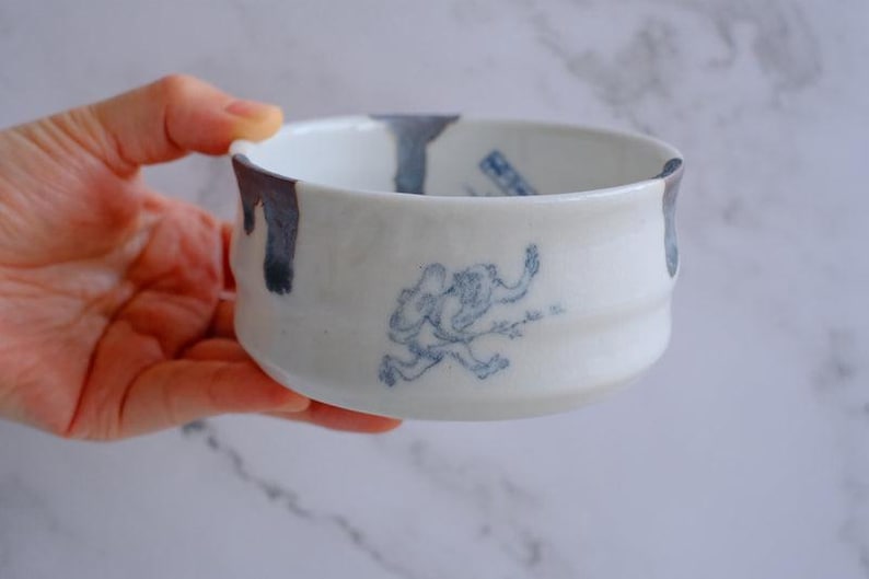 May include: White ceramic bowl with a blue illustration of a running figure. The rim has blue paint drips. The bowl has a slightly textured surface and a rounded shape.