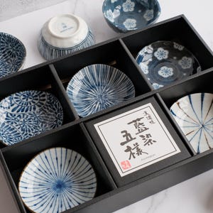 May include: A set of blue and white ceramic bowls and plates arranged in a black wooden box. The bowls and plates feature various patterns, including floral and geometric designs. A white label with Japanese text is in the center.