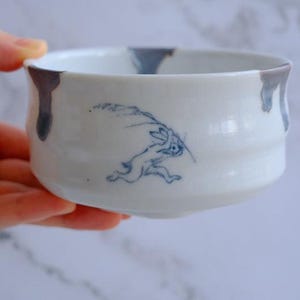 May include: White ceramic bowl with a blue rabbit illustration. The bowl has a rounded shape with a slightly flared rim. The rim is accented with dark gray drips. The rabbit is depicted in a running pose, adding a dynamic element to the design.