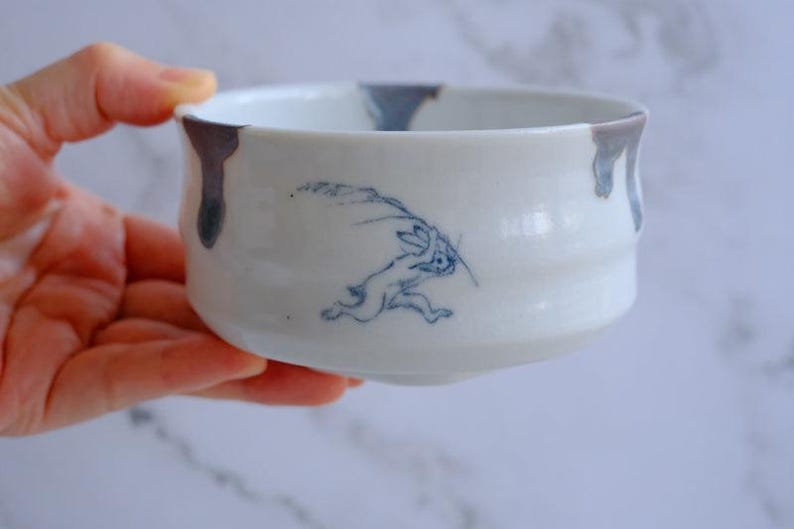 May include: White ceramic bowl with a blue rabbit illustration. The bowl has a rounded shape with a slightly flared rim. The rim is accented with dark gray drips. The rabbit is depicted in a running pose, adding a dynamic element to the design.