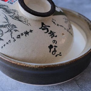 Puede incluir: Una olla japonesa de cer&aacute;mica nabemono con tapa. La tapa presenta una ilustraci&oacute;n en blanco y negro y texto japon&eacute;s. La olla es marr&oacute;n y beige con un interior blanco. La olla tiene dos asas.