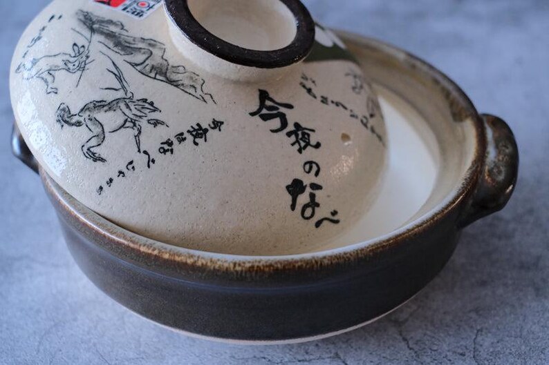 Puede incluir: Una olla japonesa de cer&aacute;mica nabemono con tapa. La tapa presenta una ilustraci&oacute;n en blanco y negro y texto japon&eacute;s. La olla es marr&oacute;n y beige con un interior blanco. La olla tiene dos asas.