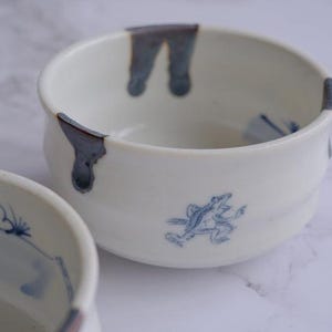 May include: Two white ceramic bowls with blue accents. One bowl has a blue frog illustration, and both have blue drips on the rim. The bowls are on a white surface.