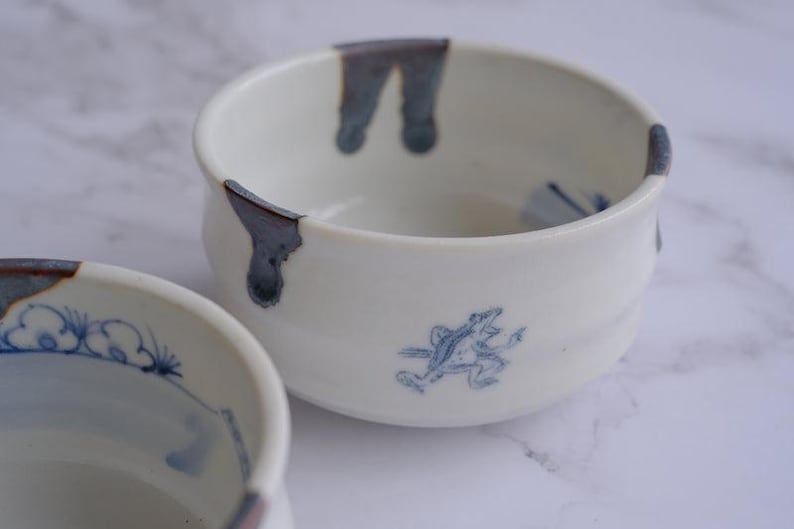 May include: Two white ceramic bowls with blue accents. One bowl has a blue frog illustration, and both have blue drips on the rim. The bowls are on a white surface.