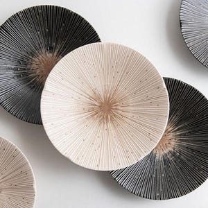 May include: A collection of decorative ceramic plates. The plates feature a radial design with a textured surface. The color palette includes black, cream, and a warm brown center. The plates are arranged on a white background.